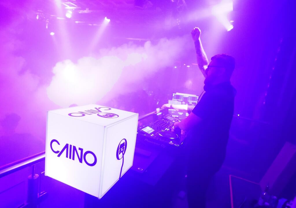 DJ Caino performing beside his branded cube under the purple nightclub lightning